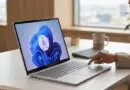 Laptop on a desk with a Windows login screen showing a fingerprint icon; a person taps the touchpad nearby.
