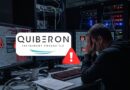 Man sits at a cluttered desk in a server room, hands on face as monitors show code and warning icons with a Quiberon logo overlay. They appear stressed by a cybersecurity alert.