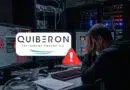 Man sits at a cluttered desk in a server room, hands on face as monitors show code and warning icons with a Quiberon logo overlay. They appear stressed by a cybersecurity alert.