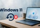 Laptop on a wooden desk displaying Windows 11 wallpaper, with a large Windows 11 text and a circular refresh icon accompanied by a red X, implying an error or failure
