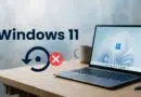 Laptop on a wooden desk displaying Windows 11 wallpaper, with a large Windows 11 text and a circular refresh icon accompanied by a red X, implying an error or failure