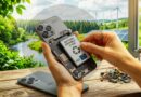 Hands replace a smartphone battery on a wooden table outdoors, with wind turbines and solar panels in the green background.