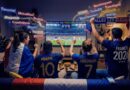 Group of France 2026 soccer fans in a living room, wearing blue jerseys and waving scarves as they cheer a match on the TV.