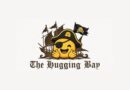 Logo featuring a smiling pirate-emoji with an eyepatch hugging its cheeks in front of a sailing ship, with the gothic text The Hugging Bay beneath it.