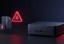 Small black desktop mini PC on a dark surface with a glowing red malware warning triangle and bug icon nearby; a circular button shows a downward arrow stuck to a case on the left.